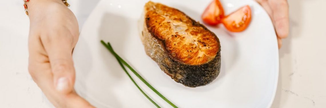 Close-up of grilled salmon served with tomatoes and chives on a plate, held by tattooed hands.