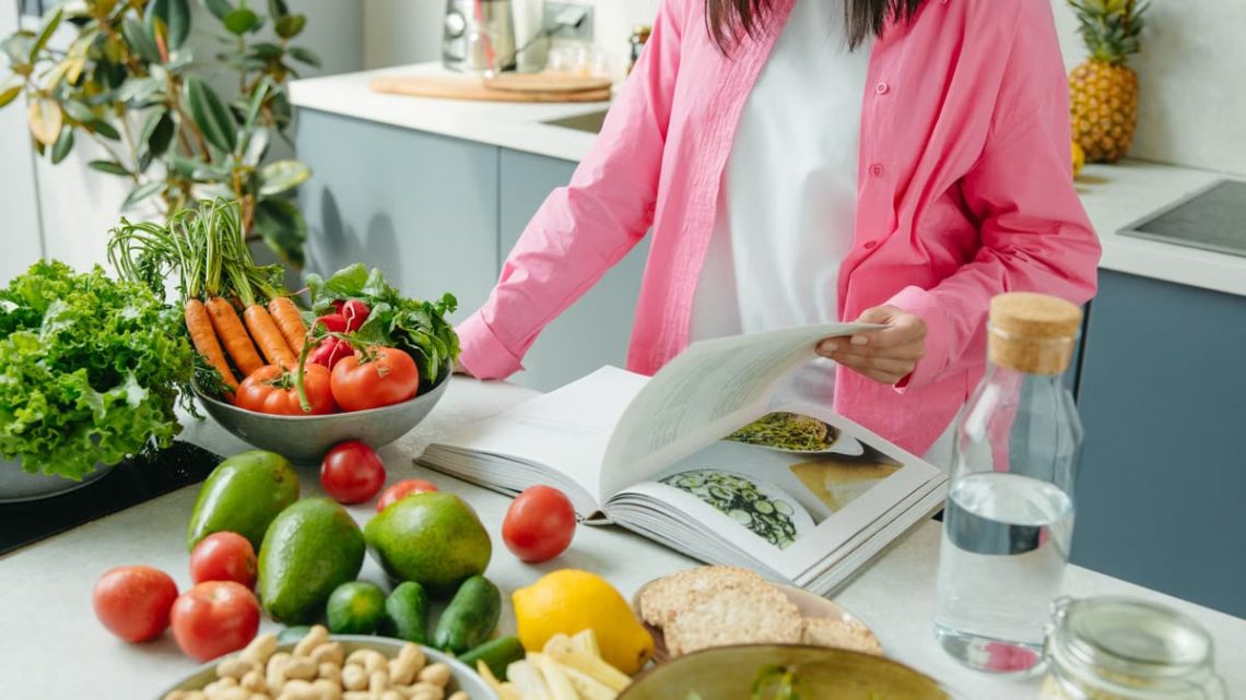 A person browsing a cookbook surrounded by fresh vegetables in a bright kitchen setting.