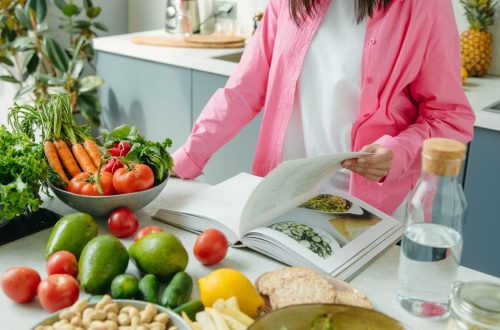 A person browsing a cookbook surrounded by fresh vegetables in a bright kitchen setting.